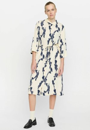 Knieumrangendes Kleid in Creme mit blauem Blumenmuster, mit Kragen, dreiviertelärmlig und mit Kordelzug in der Taille. Getragen mit schwarzen Slippern.