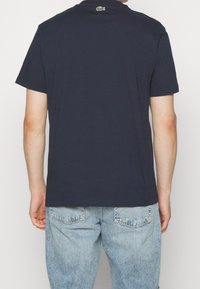 Man wearing a navy blue polo shirt with a small green crocodile logo on the upper back, paired with light blue denim jeans.