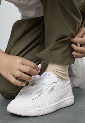 Enfant attachant une chaussure blanche Puma avec des bandes Velcro, portant des chaussettes beiges et un pantalon vert olive, assis sur un sol gris.