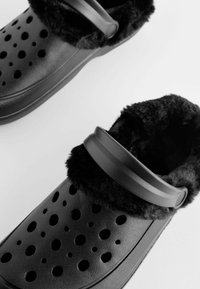 Schwarze Clogs zum Hineinschlüpfen mit einem weichen schwarzen Futter aus Fell, ausgestattet mit großen Belüftungslöchern und einem breiten Riemen über dem Spann für einen sicheren Sitz.