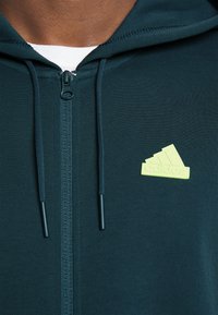 Tamnozeleni zip-up dukserica s teksturiranim završetkom, koja ima bijeli unutarnji sloj i svijetlozeleni Adidas logo na prsima.