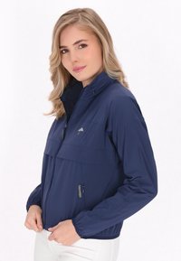 Marinblå lättviktsjacka med dragkedja fram, elastiska muddarna och en tvåfärgad design. Har en logotyp på bröstet och en sidoficka.