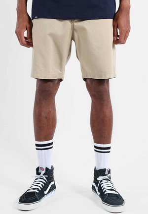 Homme portant un short beige, des chaussettes blanches montantes avec des rayures noires, et des baskets montantes noires, debout devant un fond blanc.
