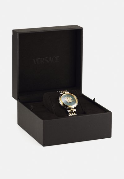Aranyszínű óra zöld számlappal, amelyen a Medusa embléma található, fekete dobozban, puha, sötét belsővel. A dobozon a "VERSACE" felirat látható.