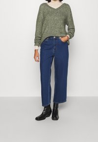 Grön stickad tröja med V-ringning, tillsammans med högmidjade blå vida jeans och svarta ankelboots. Texturerad tyg på tröjan.