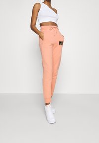 Peach sweatpants with a tapered fit, elastic waistband, and drawstring. Features side pockets and a black and gold logo patch on hip.