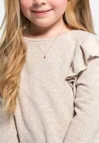 Collana d'oro con un piccolo pendente di pietra preziosa rossa, indossata sopra un maglione beige a maglia con dettagli a volant sulle maniche. Texture morbida visibile.