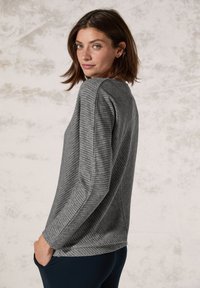 Graues Langarmshirt mit strukturiertem, geripptem Muster, ausgestattet mit einem Rundhalsausschnitt und seitlichen Taschen, kombiniert mit dunkelblauen, engen Hosen.