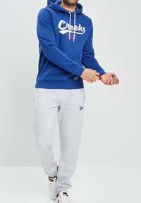 Sudadera con capucha azul y logo en blanco, combinada con pantalones de chándal gris claro. Ambos hechos de una mezcla de algodón, con cordones y bolsillos.