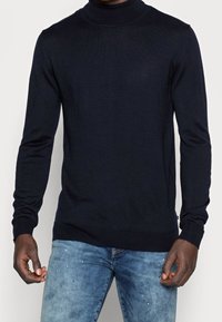 Pull-over col roulé marine en tissu côtelé avec manches longues et coupe ajustée, associé à un jean bleu clair légèrement usé.