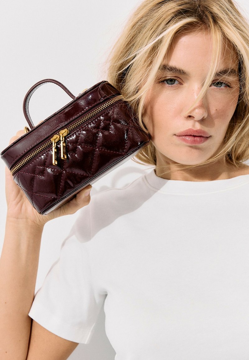 Femme blonde en chemise blanche tient un petit sac à main en cuir matelassé rouge foncé avec des fermetures éclair dorées près de son visage.