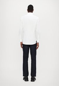 Camisa branca de manga comprida com gola, combinada com jeans de ganga escuros, exibida de trás, mostrando uma silhueta ajustada.