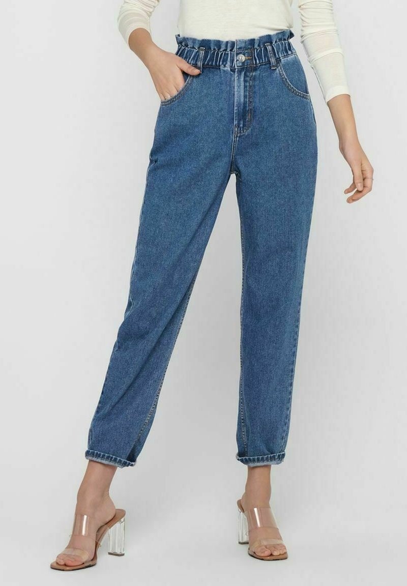 Femme portant un jean denim bleu taille haute avec une ceinture froncée et des sandales à talons hauts à bride transparente, posant avec une main dans la poche.