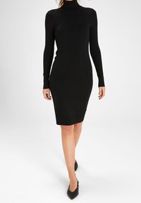 Robe noire côtelée avec un col montant et des manches longues, silhouette ajustée, longueur au genou, fabriquée en tissu tricot extensible. Chaussures noires.