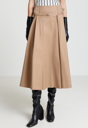 Jupe beige trapèze avec plis, ornée d'un détail métallique circulaire à la taille, assortie à des gants en cuir noir et à des bottes montantes.