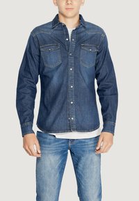 Camicetta in denim blu scuro con bottoni, due tasche frontali, cuciture a contrasto e colletto classico, abbinata a jeans blu chiaro.