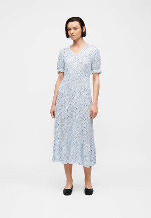 Jeune femme aux cheveux courts portant une robe midi bleu clair à fleurs avec des manches bouffantes et des ballerines noires, debout devant un fond blanc.