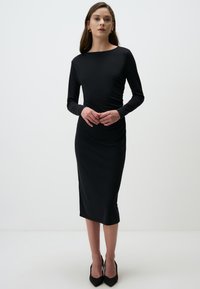 Robe noire à manches longues avec une silhouette ajust�ée, texture lisse, longueur au genou et détails de fronces sur les côtés. Assortie à des talons noirs.