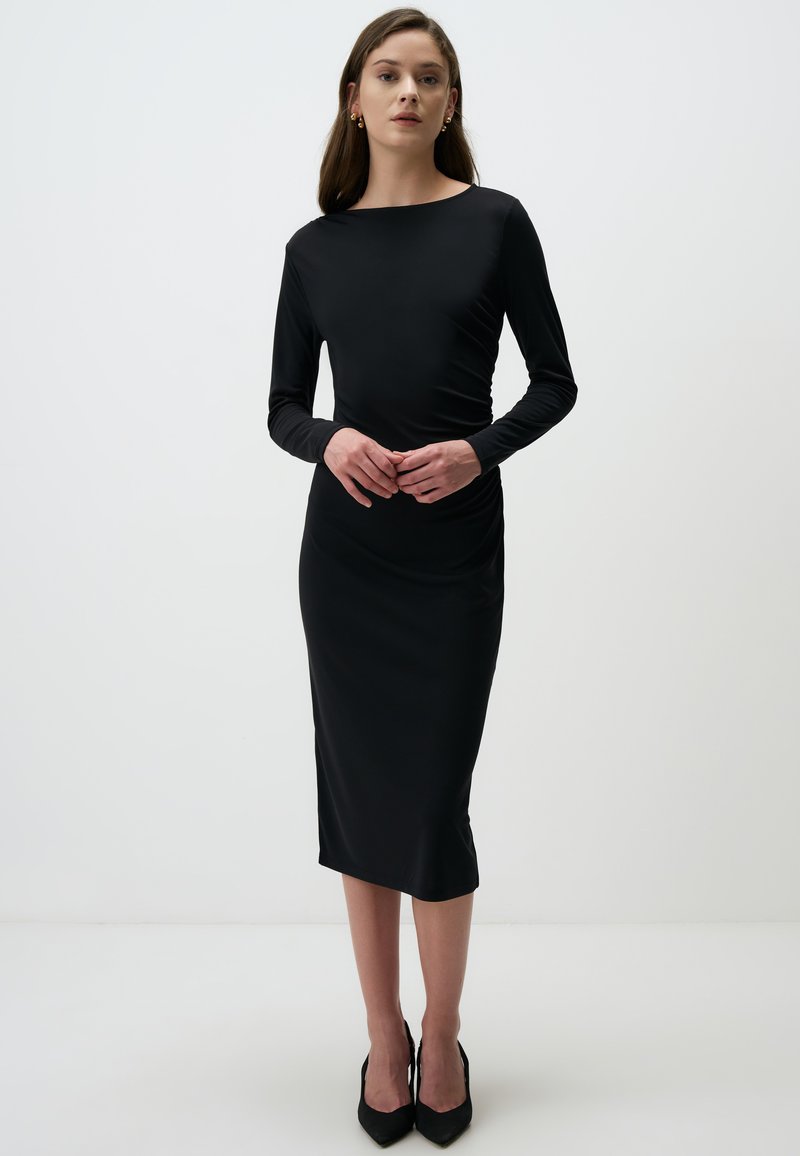 Robe noire à manches longues avec une silhouette ajustée, texture lisse, longueur au genou et détails de fronces sur les côtés. Assortie à des talons noirs.