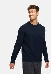 Marineblauwe gebreide trui met een ronde hals en geribde manchetten, met een gladde textuur en een iets relaxte pasvorm, gecombineerd met grijze broek.