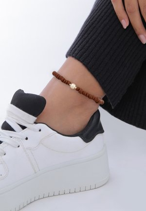 Brun perleankelarmbånd med en central guldfarvet perle, båret på en fod over en hvid sneaker med sorte accenter.