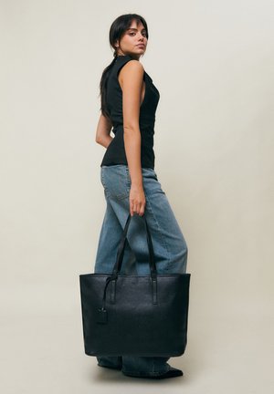 Femme debout de profil portant un haut noir sans manches et un jean bleu, tenant un grand sac fourre-tout en cuir noir par les poignées.