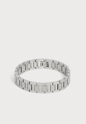 Bracelet en métal argenté avec des segments rectangulaires, certains décorés de petits cristaux transparents, posé à plat sur une surface blanche.