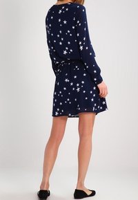 Kvinna klädd i en marinblå långärmad klänning med vita stjärnmönster, tillsammans med svarta platta skor, står med ryggen vänd mot kameran.