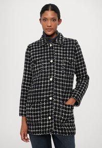 Abrigo de cuadros en blanco y negro con cuello, bolsillos frontales y cierre de botones. La tela parece texturizada y tiene una sensación suave y cálida.