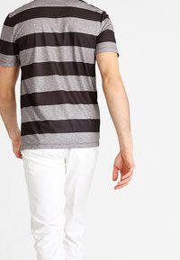 Chemise à manches courtes avec rayures grises et foncées, en tissu texturé. Portée avec un pantalon blanc, affichant un style décontracté.