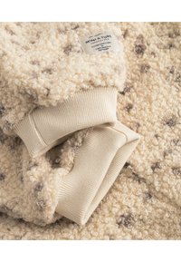 Cremefarbener Fleecepullover mit einer weichen, flauschigen Textur, ausgestattet mit einem gestrickten beigen Bündchen und einem Etikett, auf dem "MINI A.T.U.R.E." mit dunkelbraunen Punkten steht.