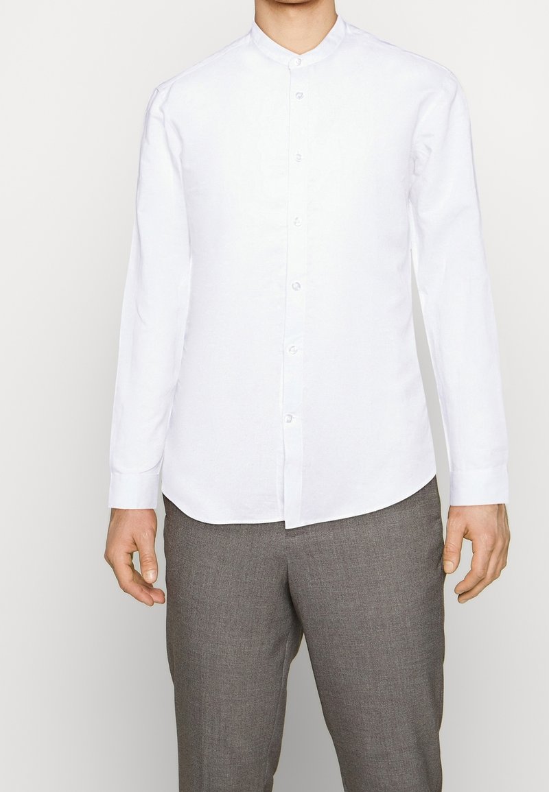 Camicia bianca a maniche lunghe con colletto a band. Realizzata in materiale leggero. Presenta una chiusura con bottoni e un orlo curvo. Texture liscia.