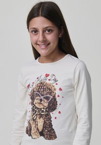 Mädchen mit langen braunen Haaren, das lächelt und ein weißes Langarmshirt mit einem Grafikmotiv von einem lockigen Hund in Sonnenbrillen und Herzen trägt.