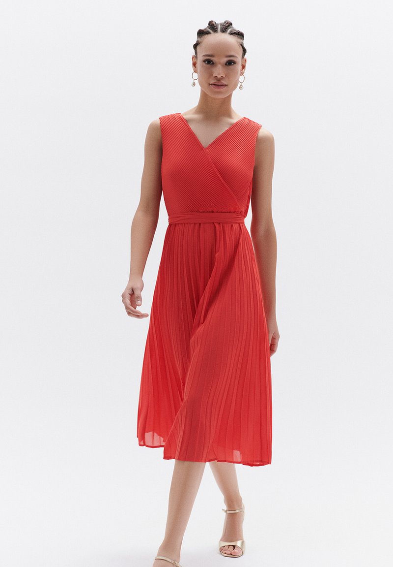 Rode, geplooide mididress met een V-hals en een ceinture taille. Glad textuur met een vloeiende silhouet, gecombineerd met beige sandalen met bandjes.