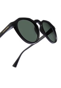 Ochelari de soare negri cu un design geometric, lentile de culoare verde închis și branding auriu pe tâmplă. Prezintă brațe subțiri negre.