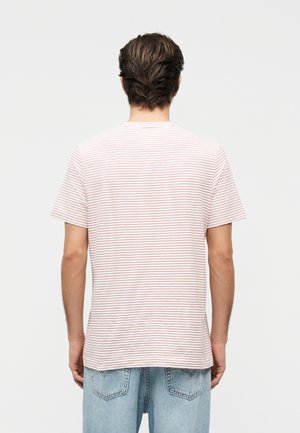 Homme aux cheveux bruns courts portant un T-shirt blanc à fines rayures horizontales rouges et un jean bleu clair, vu de dos.