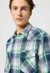Koszula w zielono-granatową kratę wykonana z bawełny, z kołnierzykiem z wyłogami, dwoma kieszeniami na piersi z zatrzaskami oraz metką z logo Wrangler.