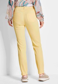 Gelbe, enganliegende Hosen mit Gesäßtaschen, taillierte Ausführung und glattes Material. Getragen mit einer gemusterten Bluse und hellen Sneakers.