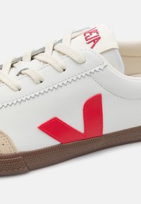 Zapatilla blanca con un logo 'V' de goma roja. Presenta un detalle de ante beige, cordones beige y una suela de goma marrón texturizada. Acabado de cuero suave.
