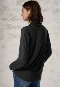 Haut à manches longues noir avec une texture douce et un col froncé. Porté avec un jean bleu, affichant une coupe décontractée et une silhouette lisse.