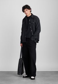 Svart knapp-up skjorta jacka över en mörk tröja, kombinerat med svarta lösa byxor och svarta och vita skor. Håller en svart tote-väska.