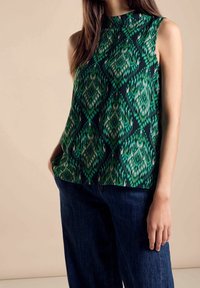 Ärm-lös topp i ett grönt och svart mönster, med hög halsringning och lös passform, i kombination med mörkblå vida jeans.