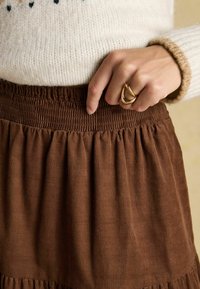 Brun manchesterkjol med samlad struktur och elastisk midjeband, kombinerad med en crèmefärgad tröja och en guldring på handen.