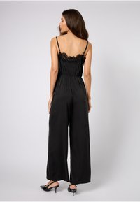 Svart jumpsuit med blonder detaljer på ryggen. Har tynne stropper, innsvinget kropp og vide ben. Glatt tekstur og elegant design.