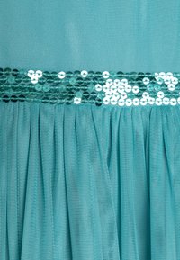 Tyrkysová tylová látka s vrstveným designem, obsahující horizontální pás lesklých tyrkysových flitrů v pase pro zvýraznění textury a detailu.