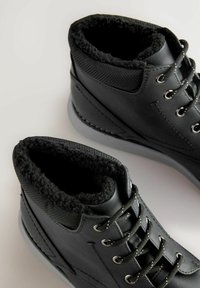 Botas negras por el tobillo con un exterior de cuero suave, forro de tela texturizada y un suave ribete de piel alrededor del cuello; cuenta con ojales metálicos y suelas grises.