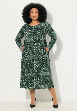 Grünes und schwarzes gemustertes langärmliges Kleid mit Rundhalsausschnitt, Taschen und fließender Silhouette, kombiniert mit schwarzen Heels.