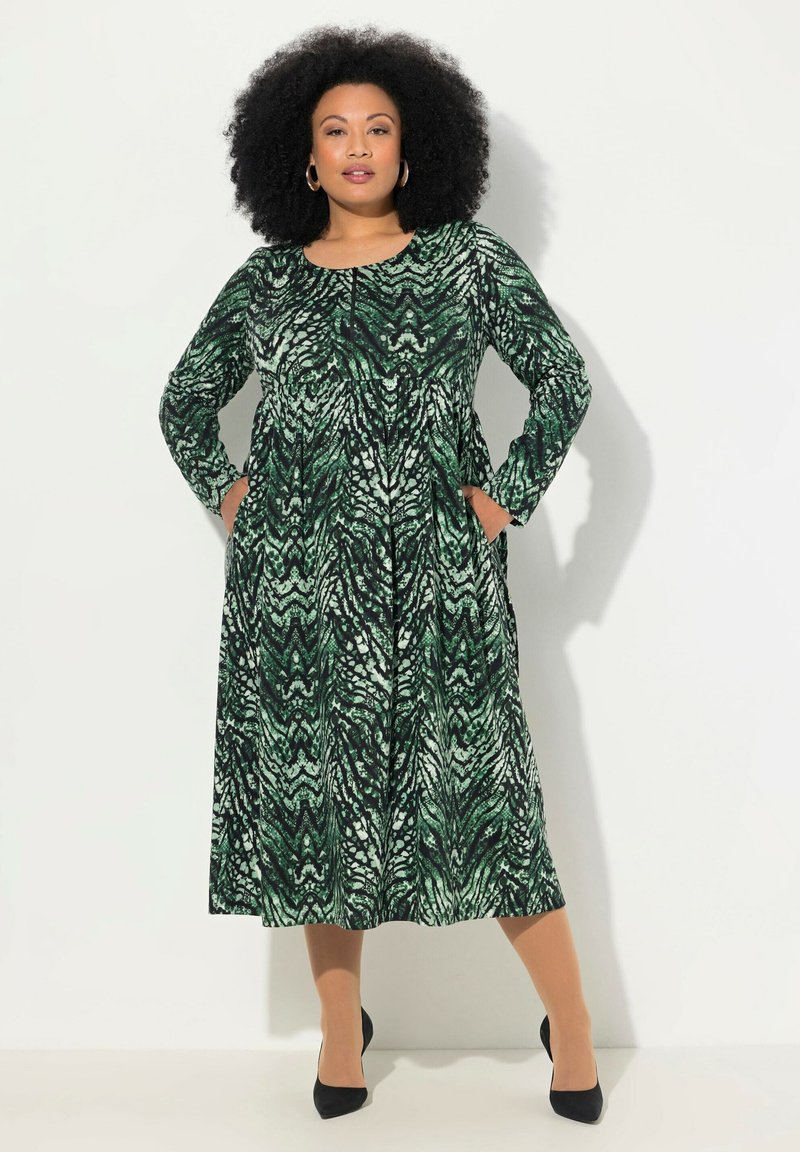 Vihreä ja musta kuviollinen pitkähiha dressi, jossa on pyöreä kaula-aukko, taskut ja väljä siluetti, yhdistettynä mustiin korkokenkiin.