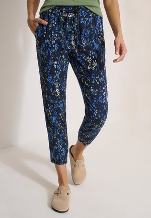 Personne portant un pantalon court bleu marine avec des motifs floraux bleus et verts, des chaussures beiges à enfiler et un haut vert, debout avec une main dans la poche.
