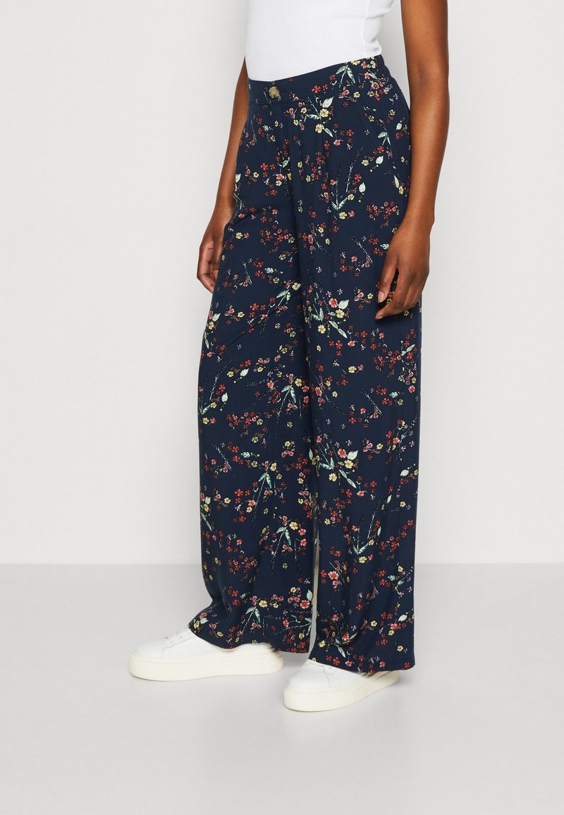 Personne portant un pantalon large bleu marine avec un petit imprimé floral multicolore, des baskets blanches et un haut blanc, debout devant un fond uni.